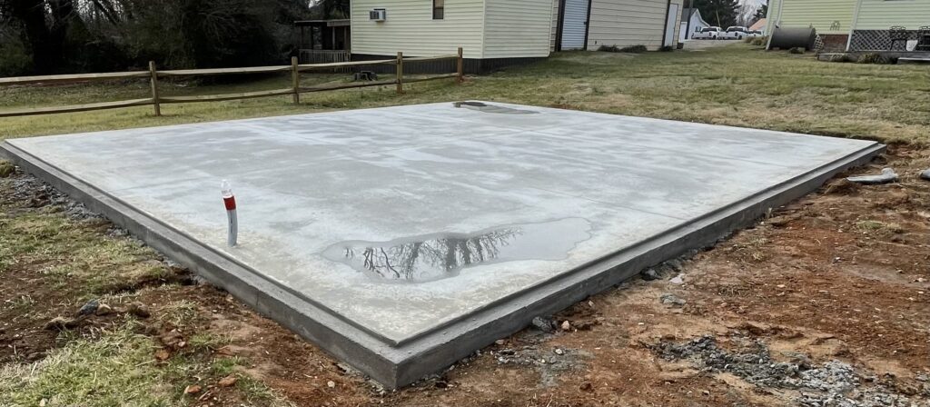 Finished concrete slab foundation with raised perimeter footing, shown in a residential yard with fresh curing marks and a plumbing stub-out visible near the corner.